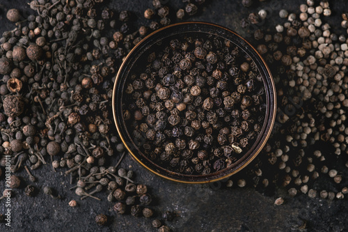 Variety of different black peppers allspice, pimento, monks pepper, peppercorns in tin can over old black iron texture background. Top view, space. Close up