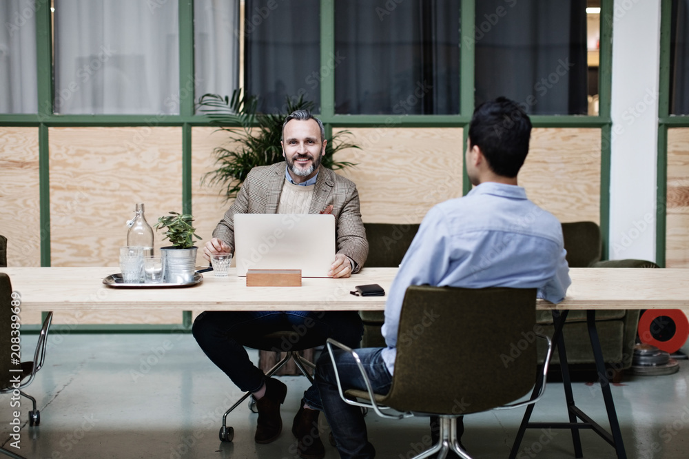 Businessman using laptop while sitting with colleague at table in office Businessman using laptop while sitting with colleague at table in office