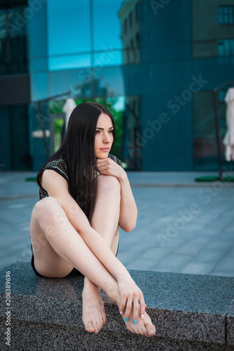 portrait of young girl with long brunette hair, sitting on the curb near skyscraper, girl wearing brown blouse and black skirt