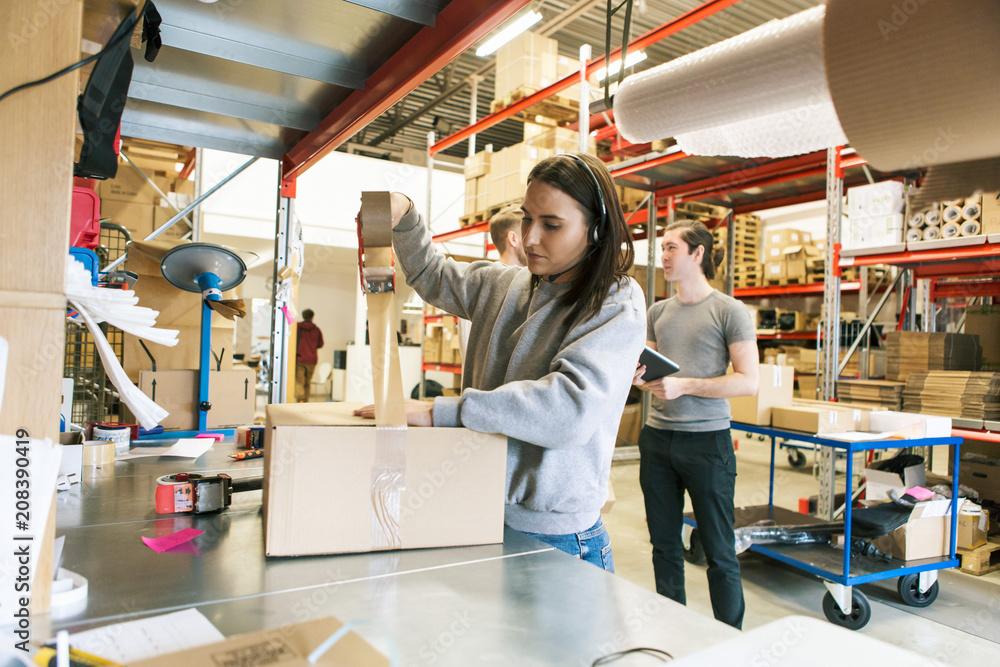 Woman packing box while coworkers discussing in distribution warehouse Woman packing box while coworkers discussing in distribution warehouse