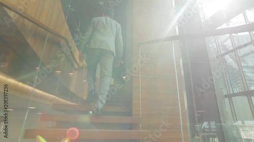 Businessman going up stairs back low angle view modern wooden glass interior commodious house. Man wearing business clothes suit ascending climbing upstairs in office apartment. Building design style photo