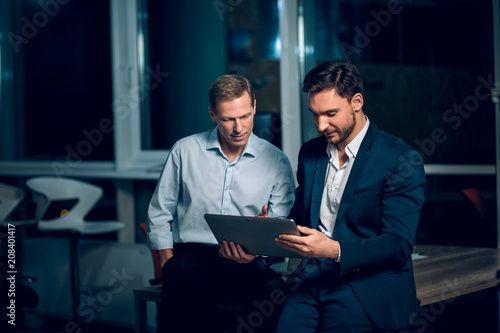 Two office collegues looking at tablet. Handsome businessman wearing suit showing something to his collegue on tablet late in evening.