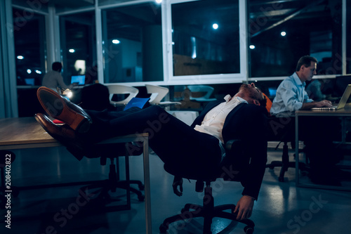 Tired man sleeping in office after overworking. Exhausted businessman sleeping in his office chair with his legs up on office table in middle of night.