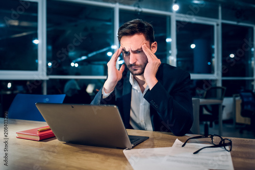 Weary businessman finishing up on work at night. Stressed and tired office worker rubbing his forehead with both of his hands while working behind computer in late hours in office.