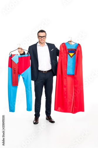 businessman in eyeglasses holding hangers with superhero costume and smiling at camera isolated on white