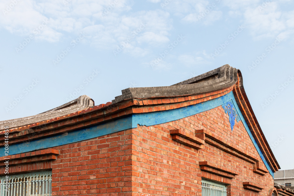 Traditional swallowtail style roof line found in historic villages in China and Taiwan. The building is brick with a decorative blue symbol & band around the building. There is a blue sky and clouds. Traditional swallowtail style roof line found in historic villages in China and Taiwan. The building is brick with a decorative blue symbol & band around the building. There is a blue sky and clouds.