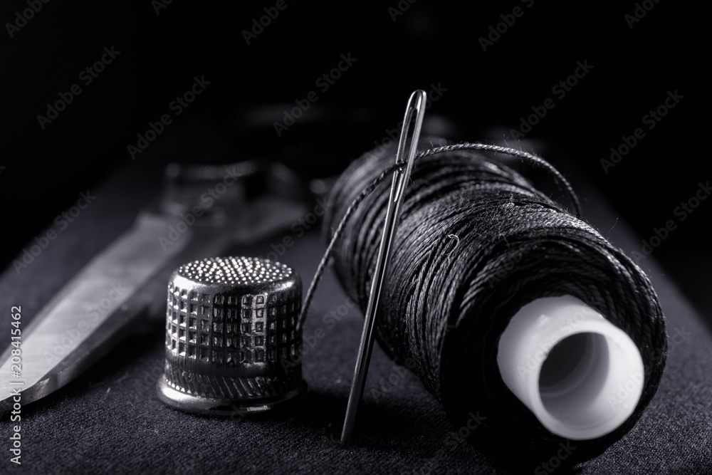 Needle with thread, thimble, bobbin with black thread and scissors on black  background Stock Photo