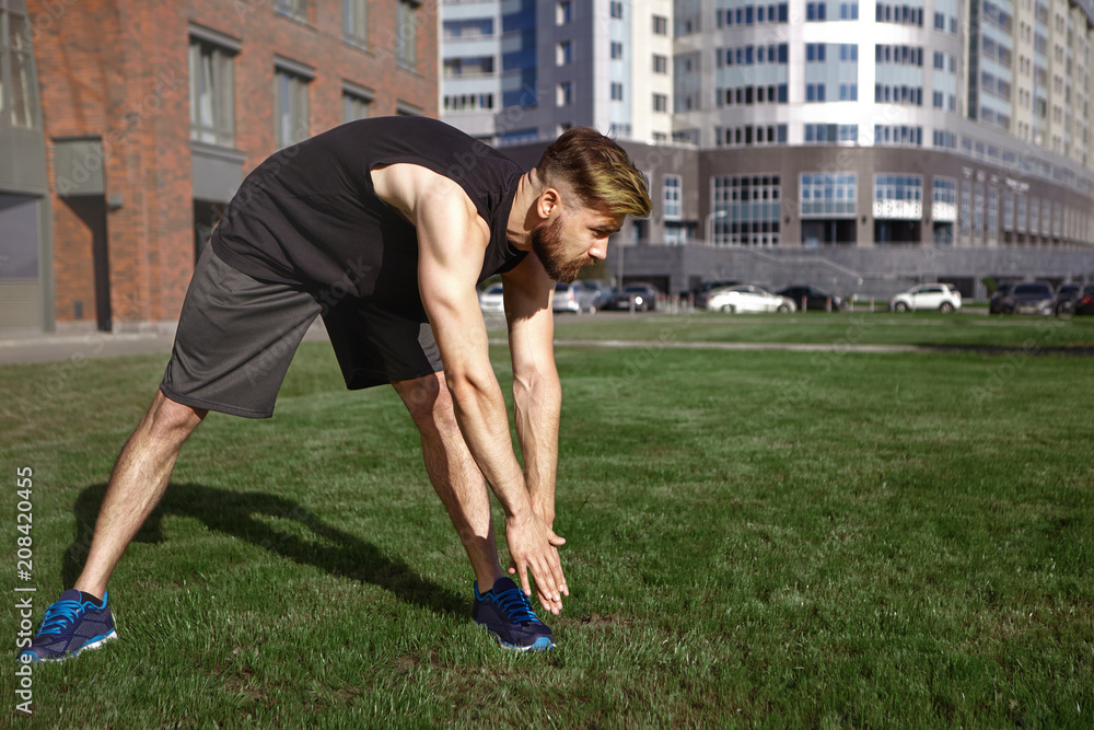 Fototapeta premium Summertime, activity, fitness and hobby concept. Muscular young European unshaven man in black sports clothes and running shoes bending over to left toe, stretching legs while exercising outdoors