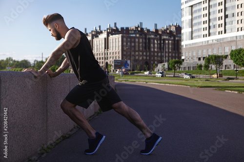 Sideways outdoor picture of handsome young male jogger with stubble and muscular athletic body doing stretching exercise on sidewalk, preparing for running morning workout, having confident look