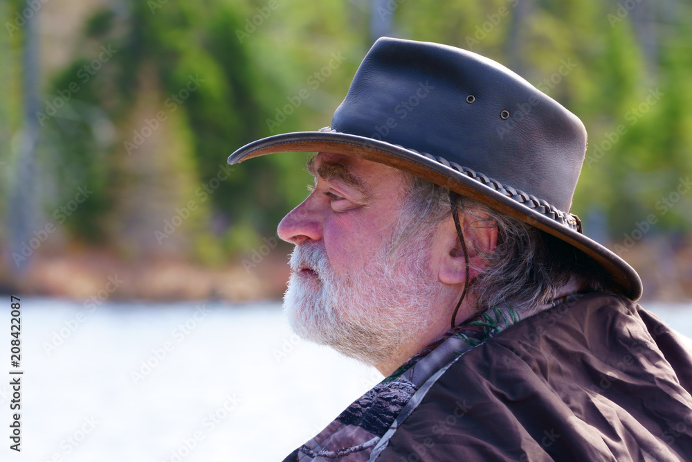 A peaceful caucasian man, 50's, wearing a white beard, a leather hat Australian style and a camouflage coat, is fishing in his boat on a lake with blue water by a beautiful, fresh, sunny morning in th