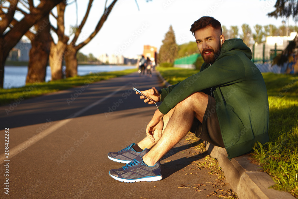 Obraz premium Street style, fashion and electronic devices. Happy smiling young man with stubble listening to audiobook using headphones and mobile phone, enjoying high speed 4g internet connection during walk
