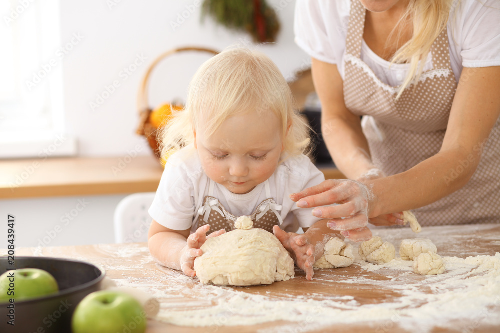Little girl and her blonde mom in beige aprons playing and laughing while kneading the dough in the kitchen. Homemade pastry for bread, pizza or bake cookies Little girl and her blonde mom in beige aprons playing and laughing while kneading the dough in the kitchen. Homemade pastry for bread, pizza or bake cookies