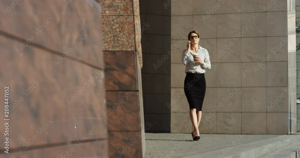 Successful attractive businesswoman in the glasses having a business talk on the phone while walking outside the big modern building and drinking coffee to go. Successful attractive businesswoman in the glasses having a business talk on the phone while walking outside the big modern building and drinking coffee to go.