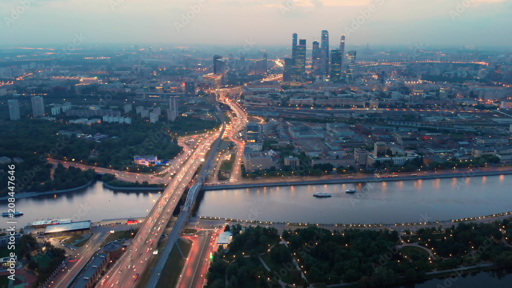 Fototapeta premium Flying above the wide illuminated city highways and Moskva river. Cityscape at dusk. Moscow-city towers in the evening haze on the horizon.