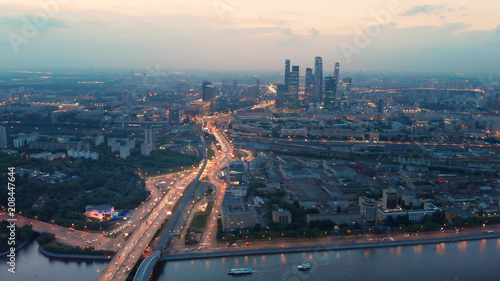 Flying above the wide illuminated city highways and Moskva river. Cityscape at dusk. Moscow-city towers in the evening haze on the horizon.