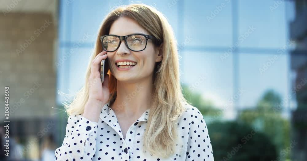 Close up of the charming woman in the business style standing near the modern building while talking cheerfully on the phone and laughing. Outdoor. Close up of the charming woman in the business style standing near the modern building while talking cheerfully on the phone and laughing. Outdoor.