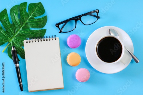 Flat lay photo with notebook , coffee cup Pen, glasses ,Macaroon and Tropical Leave on blue background