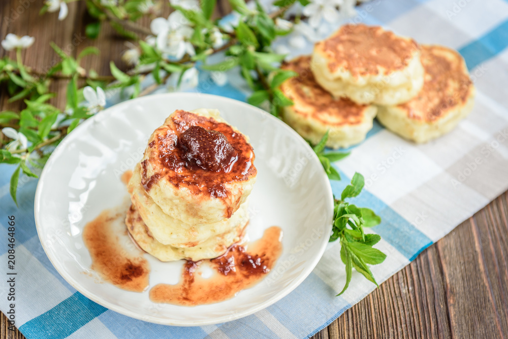 Homemade cottage cheese pancakes with strawberry jam on white plate on dark wooden background with flowers