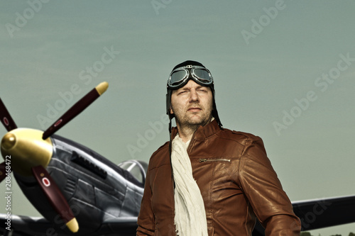 Portrait of a vintage pilot with leather cap, scarf and aviator glasses in front of a historic airplane - Portrait of a man in historical pilot clothing © Riko Best