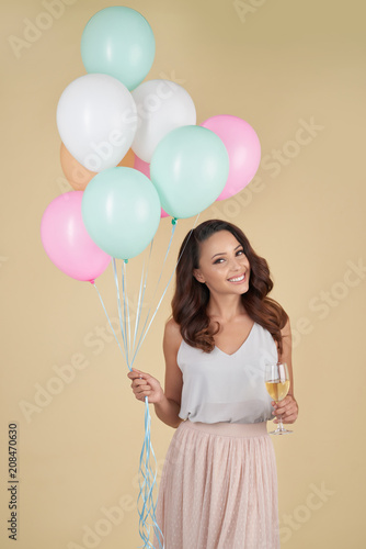 Cheerful excited attractive young woman with perfect brown hair wearing top and tender skirt holding bunch of helium balloons and champagne flute and looking at camera.