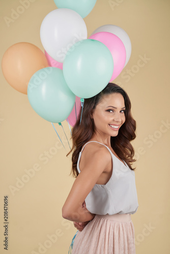 Positive excited pretty young lady in stylish top and skirt holding bunch of helium balloons behind back and looking at camera