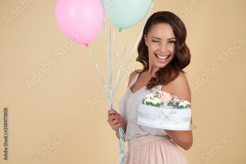 Cheerful emotional attractive young woman wearing outfit in pastel colors holding bunch of balloons and birthday cake with flowers while winking at camera against beige background.