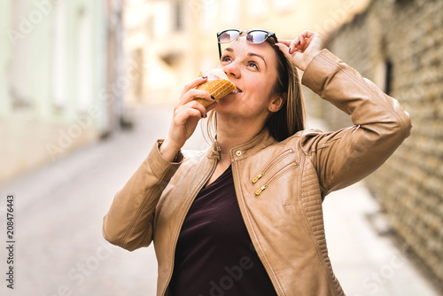 Woman lifting sunglasses and looking up while eating ice cream in city street. Tourist eating sweet dessert during travel.