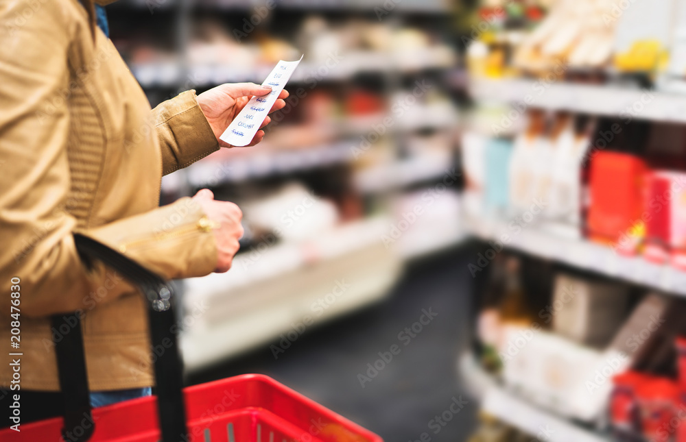 Reading shopping list in supermarket. Female customer in grocery store with  budget, plan or checklist. Lady doing groceries and buying food for family.  Shopper with basket between shelves in aisle. Stock-Foto