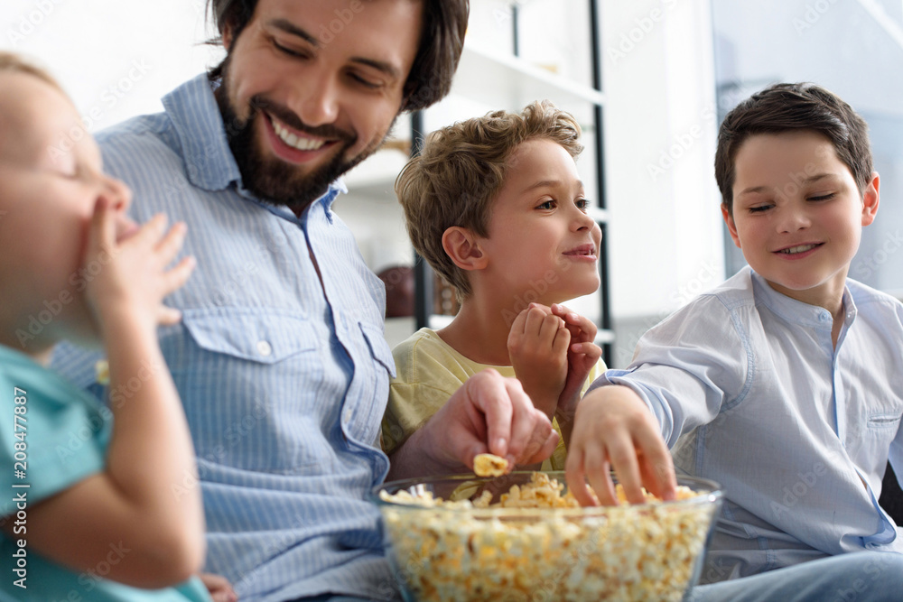 smiling man and little sons eating popcorn while watching film together at home