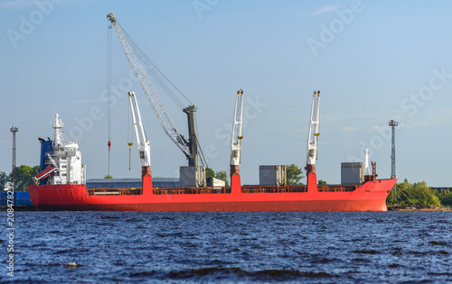 The orange cargo ship is in a river port. The crane is unloading a cement slab from the vessel.