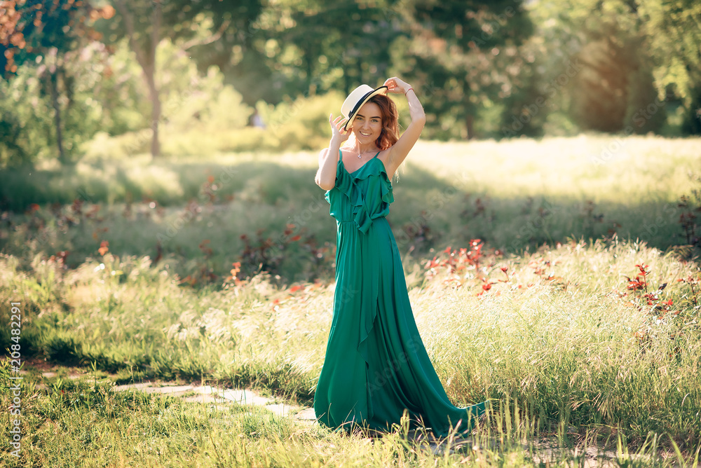 A young model girl in a long green dress and a beautiful hat walks along a path in a summer blooming garden in the background of a bright sun

