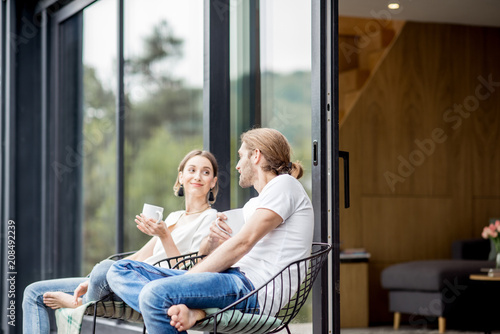 Young couple sitting with cups on the terrace of the modern house talking together outdoors