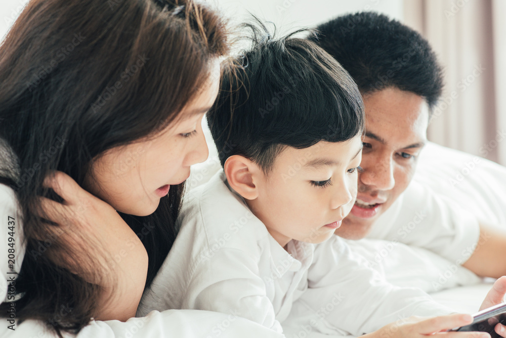 Asian boy playing smartphone with young parents lying in bed.Happy famility Asian boy playing smartphone with young parents lying in bed.Happy famility