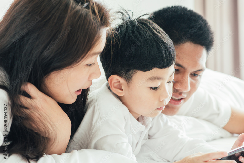 Asian boy playing smartphone with young parents lying in bed.Happy famility