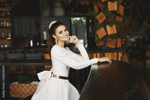 Young elegant beautiful and sexy brunette model woman in white dress with gold belt sitting in bar and waiting for boyfriend © innarevyako