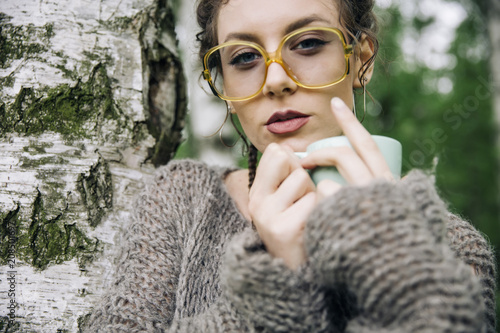 Young woman in a sweater and with glasses drinking coffee in the park