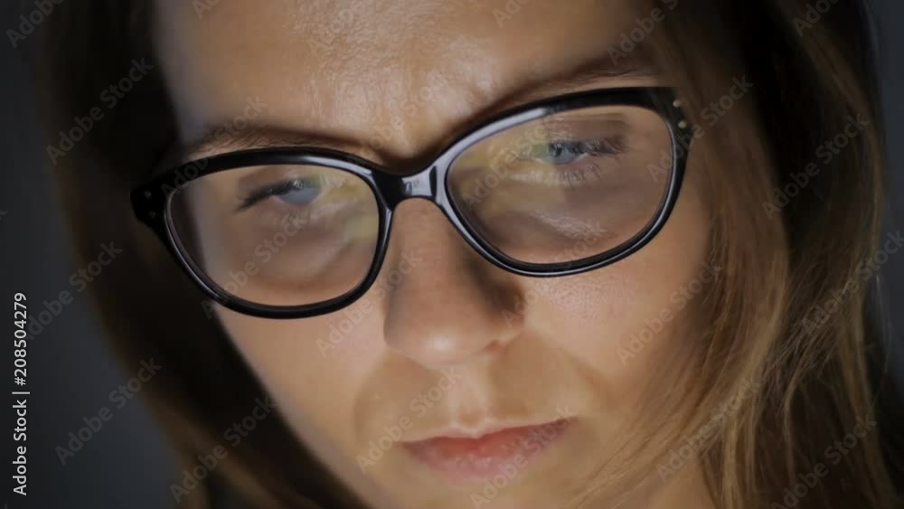 Woman is browsing news or social information feed online with her laptop, smartphone or tablet device. Monitor screen reflections could be seen in her eyeglasses glass. Woman is browsing news or social information feed online with her laptop, smartphone or tablet device. Monitor screen reflections could be seen in her eyeglasses glass.