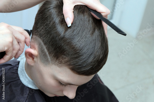The female hand of a professional hairdresser keeps a trimmer and shaves the line on the head of a teenager in a beauty salon. A black comb is clamped in the fingers of a girl with a manicure on nails