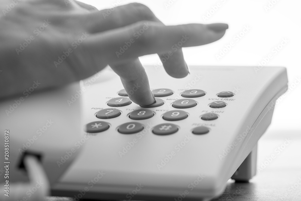 Monochrome image of female hand dialing a telephone number on a white landline phone Monochrome image of female hand dialing a telephone number on a white landline phone