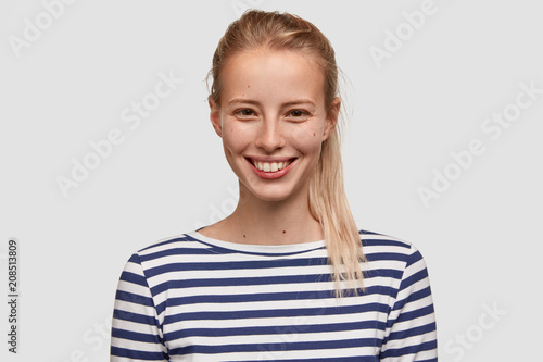 Pretty female with positive smile, wears sailor jacket, looks in satisfaction, greets new visitor in beauty salon, poses indoor against white background. Lovely glad woman rejoices her success. © wayhome.studio 