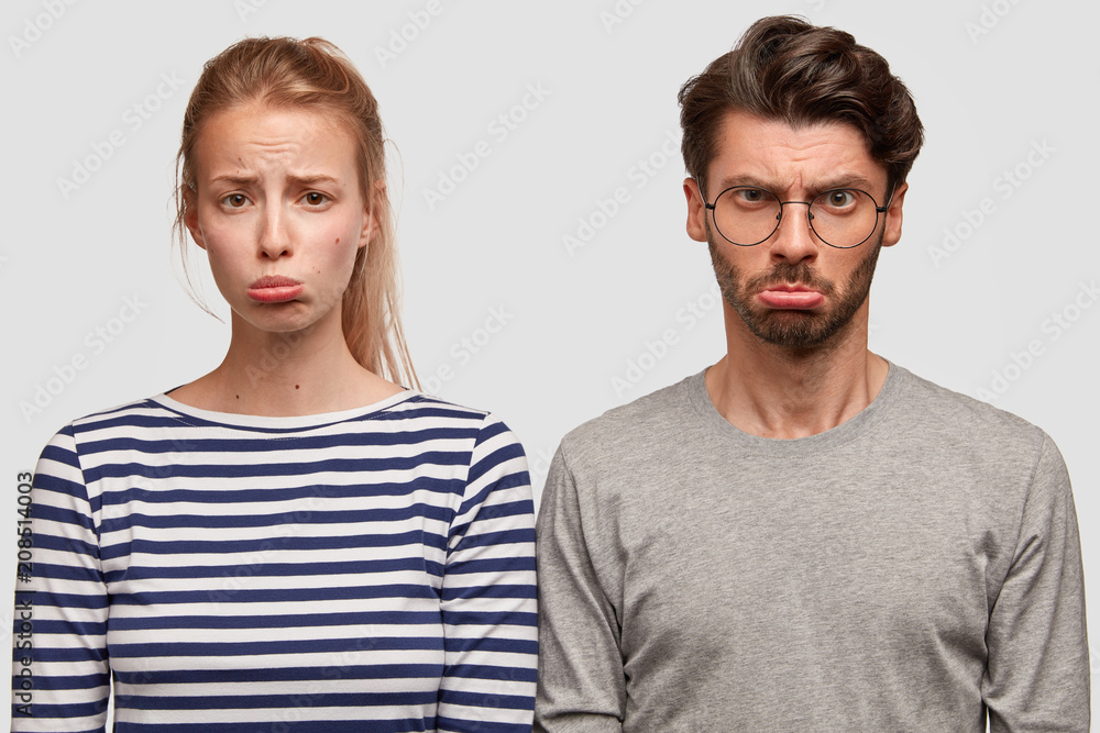 Two best friends have unhappy expressions, sullen look, stand against white studio background. Discontent European woman in striped sweater purses lower lip. Upset bearded hipster in spectacles Two best friends have unhappy expressions, sullen look, stand against white studio background. Discontent European woman in striped sweater purses lower lip. Upset bearded hipster in spectacles