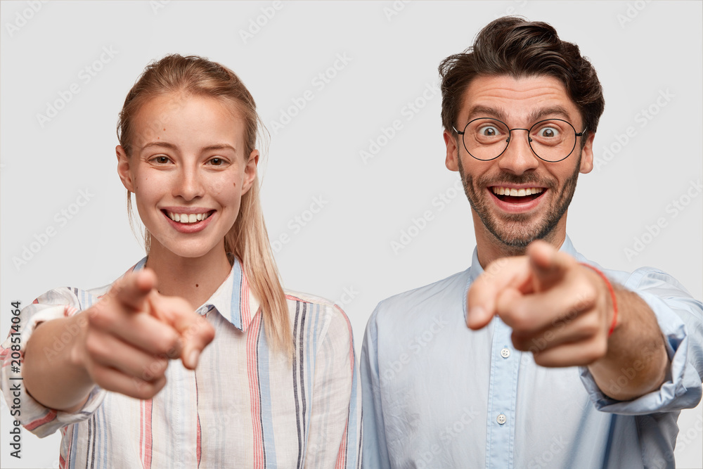 Positive friendly young woman and man with broad smiles indicate directly at camera with fore fingers, select you in their company, stand next to each other, isolated over white concrete wall. Positive friendly young woman and man with broad smiles indicate directly at camera with fore fingers, select you in their company, stand next to each other, isolated over white concrete wall.