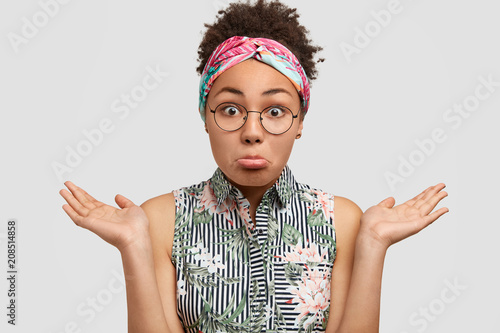 Beautiful hesitant African American female curves lower lip, shrugs shoulders, wears blouse and headband, feels doubtful as doesn`t know where to go with boyfriend, isolated over white background