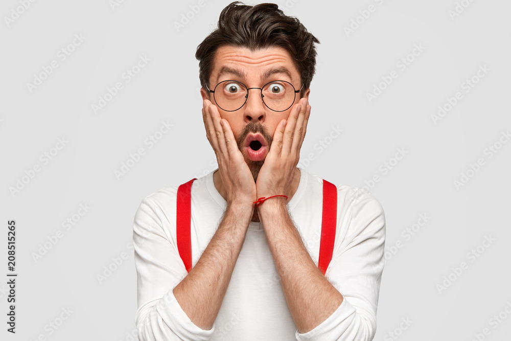 Shocked amazed male has stupefied expression, holds head with hands, stares with eyes full of disbelief, recieves unexpected task from someone, wears fashionable white shirt, poses in studio Shocked amazed male has stupefied expression, holds head with hands, stares with eyes full of disbelief, recieves unexpected task from someone, wears fashionable white shirt, poses in studio