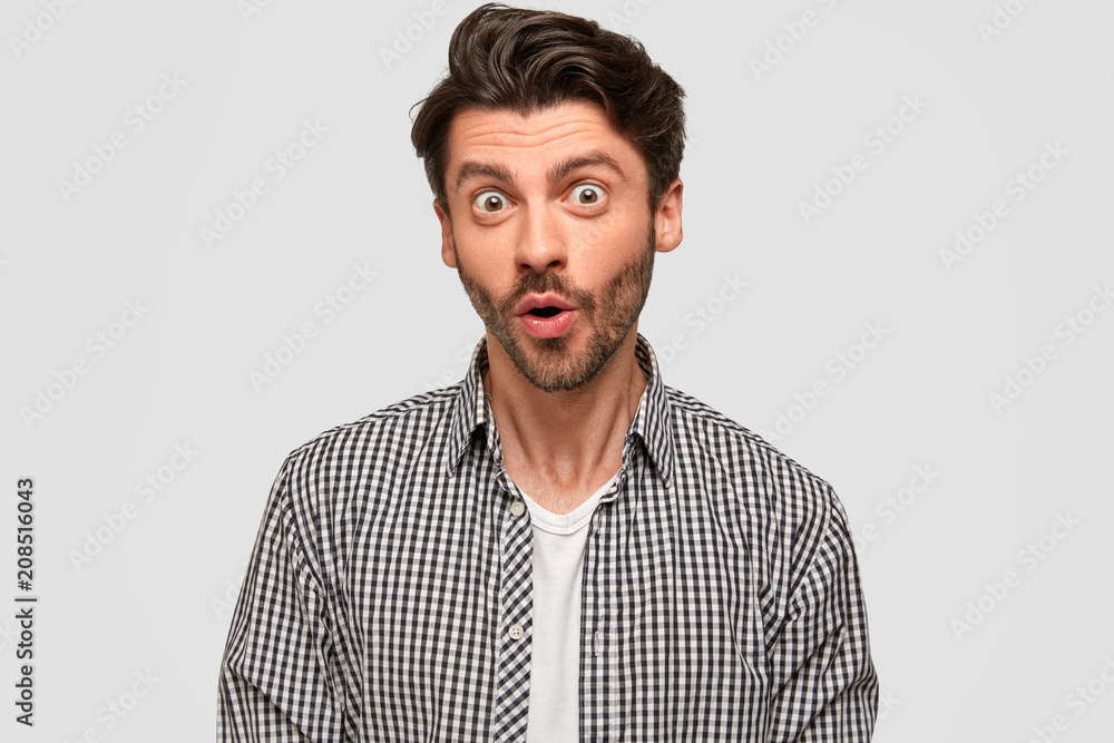 Amazed handsome male with dark hair, stubble, wears checkered shirt, eyes popped out, looks in surprisement, keeps mouth slightly opened, wonders about latest news, isolated on white studio wall Amazed handsome male with dark hair, stubble, wears checkered shirt, eyes popped out, looks in surprisement, keeps mouth slightly opened, wonders about latest news, isolated on white studio wall