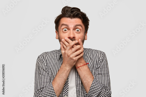 Photo of frightened Caucasian male closes mouth and stares with eyes popped out, happy to recieve proposal about job abroad, has trnedy hairstyle, isolated over white background. Emotions concept photo