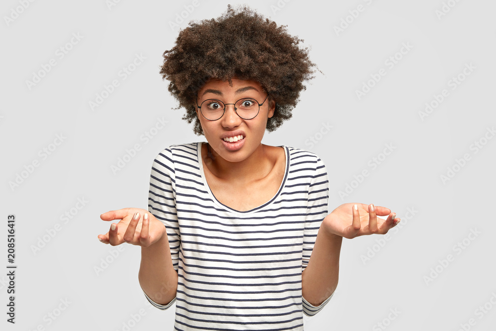 Hesitant African American female models in studio, has puzzled expression, shruggs shoulders with uncertainty, frowns face. Doubtful woman costumer can`t make choice, isolated on white wall. Hesitant African American female models in studio, has puzzled expression, shruggs shoulders with uncertainty, frowns face. Doubtful woman costumer can`t make choice, isolated on white wall.