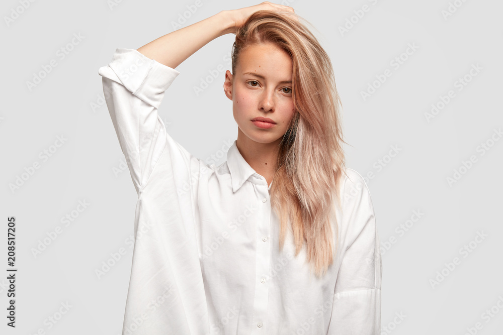 Pleasant looking gorgeous female model with long fair hair combed on one  side, wears man`s white shirt, has pure skin, keeps hand behind head, shows  natural beauty, isolated on white background Stock