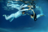 Bride and groom in a white wedding dress swim to each other underwater in the pool. Underwater wedding