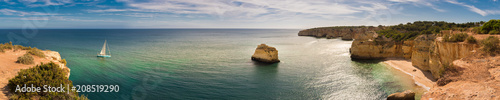Panorama of the Algarve coastline in Portugal with a sailing boat moving towards the Marinha beach. There are cliffs, vegetation, a beach and rock stacks in the scene.
